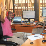A man sitting at a desk with several computer screens doing an online course. He has his hands raised in the air and is laughing at the camera.