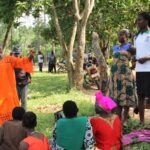 Students from African Rural University, Uganda, holding up posters and explaining things to community members outside on the grass